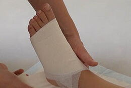 Verband Unterschenkel Zuerst wird ein ca. 10 cm langes Stück Tape längs v-förmig eingerissen. Die Basis des V wird am Fußrücken festgeklebt, die beiden 1 cm breiten Schenkel werden zur Fußsohle geführt.
